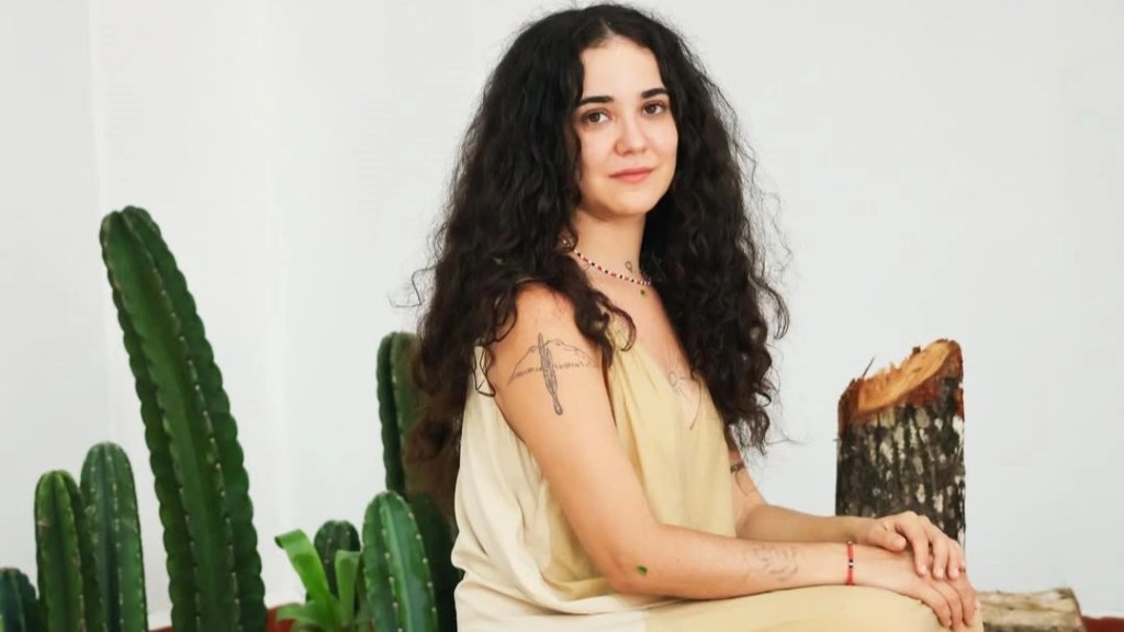 Yasmin Formiga, uma mulher de cabelos escuros e longos, sentada. Em sua volta, há cactos e um tronco de madeira cortada, representando a caatinga