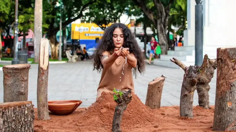 Yasmin Formiga performando ao jogar água sobre areia, com pedaços de troncos em volta e uma bacia de barro